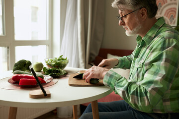 Side view of elderly man watching video recipe tutorial on smartphone to make salad being on...