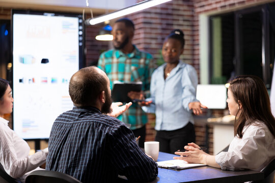 Multiethnic coworkers work efficiently in tracking progress on financial report and completing project plans. African american employees doing business presentation in late night group meeting.