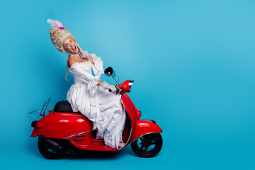 Charming woman in elegant retro costume poses on a red vintage scooter against a blue background,...