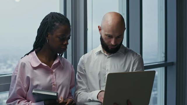 Medium portrait of diverse colleagues discussing report on wireless laptop and looking at camera at modern office workspace