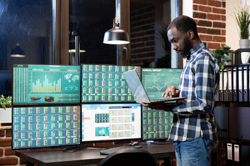 Male financial analyst with a laptop, engaged in forex trading at night, reviewing currency exchange charts. Black man standing and analyzing investment data on multiple screens in modern workstation.