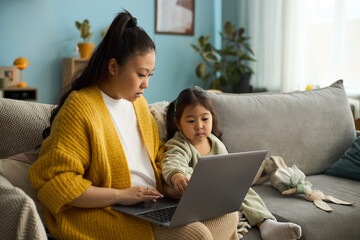 Fototapeta premium Caucasian mother and young daughter sitting on couch, using laptop together in cozy home environment, both focused and engaged without dot at the end