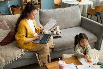 Woman in yellow cardigan sitting on couch reviewing documents with laptop on lap, while young...