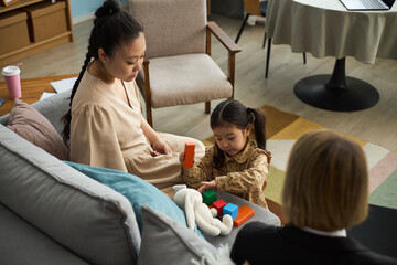 Woman sitting on couch, observing child playing with blocks, young mother focusing on activity in...