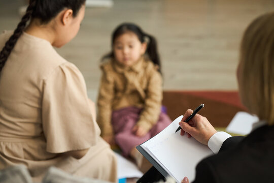 Two women discussing childs progress in kindergarten education with child present in blurred background, adult taking notes during conversation with family