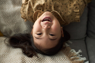 Portrait of young girl with long dark hair smiling, while lying upside down on soft couch with...