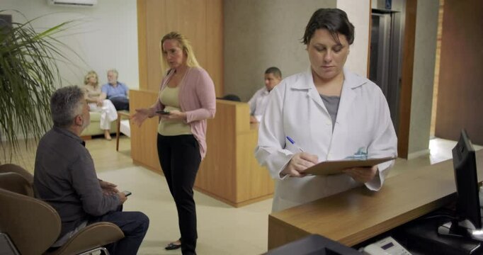 Female doctor reviewing clipboard notes at clinic reception desk while patients wait in background and staff engage with visitors in busy medical lobby