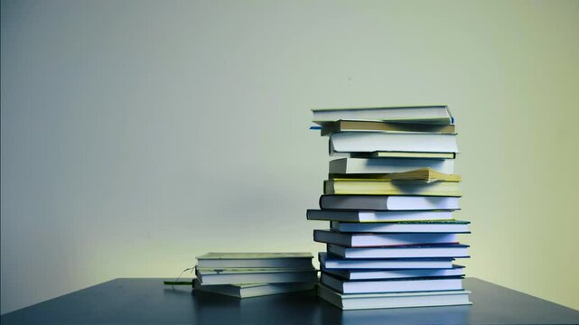 Stop motion footage of books piling up on top of each other frame by frame in timelapse mode on a table. Books are hardcover with white, yellow pages. Pile increases and then decreases on the table.
