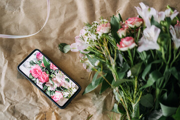 Smartphone displaying a bouquet photo next to real pink flowers on a kraft paper background. E-commerce floristry, digital product showcase, florist branding, online flower business