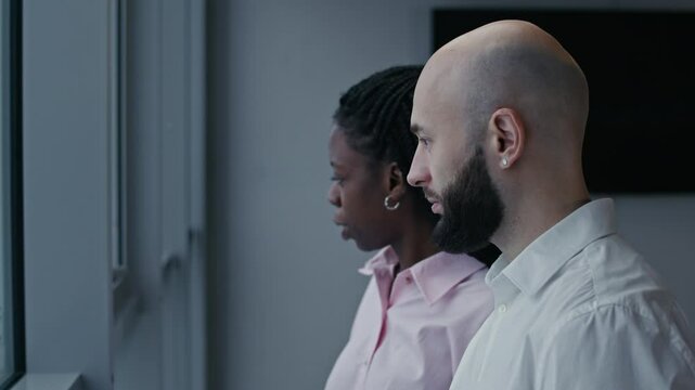 Medium shot of bicultural colleagues in formalwear having discussion by panoramic windows at modern office