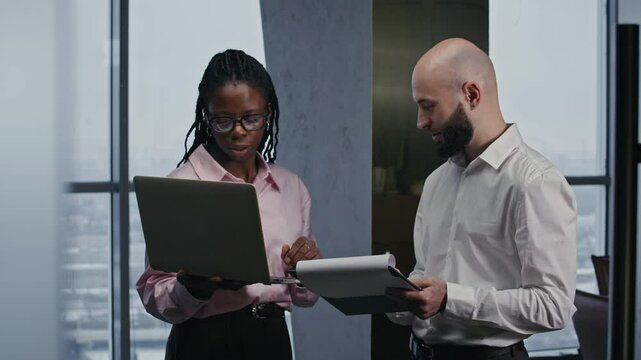 Pan shot of two intercultural people holding wireless laptop and document folder discussing business at office