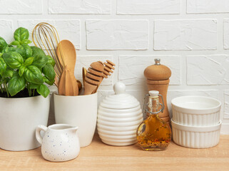 Kitchen utensils, basil plant and condiments resting on wooden countertop in front of white brick wall