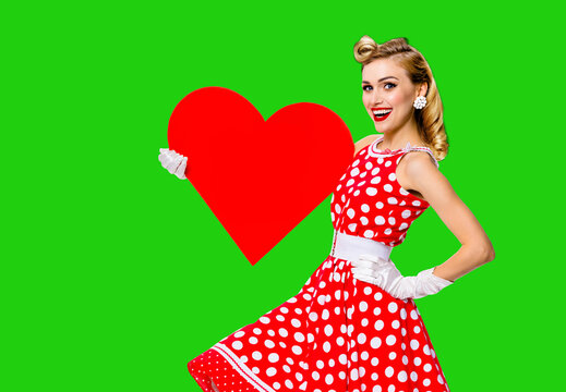 Pinup beautiful woman show red heart shape paper banner with mockup space. Pin up cute girl. Retro fashion vintage ad concept. Isolated over vibrant green chroma key background. Valentine or like sign