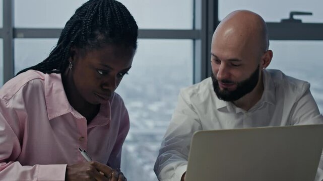 Handheld shot of bicultural employees working at desk with wireless laptop in office with panoramic windows