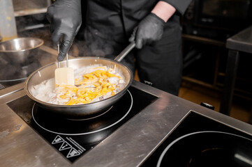 A chef in black gloves stirs creamy pasta in a frying pan on an induction stove. The professional kitchen setting is clean, emphasizing culinary expertise and delicious food preparation.