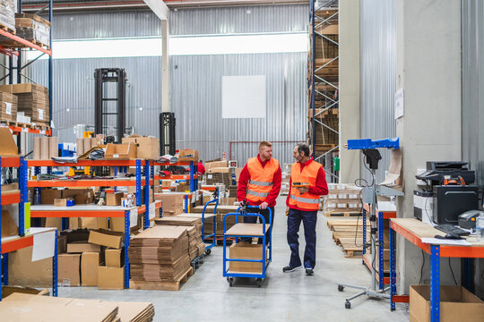 Two warehouse workers in safety vests are transporting goods on a trolley while discussing logistics in a busy warehouse environment