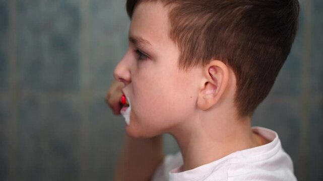 A ten year old boy is concentrating on brushing his teeth with a red toothbrush