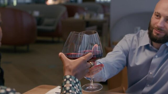 Handheld shot of bicultural man and woman clinking glasses with red wine during date in modern restaurant