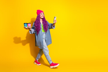 Trendy woman with vibrant pink hair and casual outfit strolling with coffee and phone against a bright yellow background.