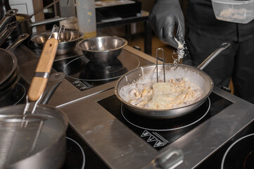 A chef wearing black gloves sprinkles grated cheese on creamy pasta in a stainless steel pan on an induction cooktop in a professional kitchen.