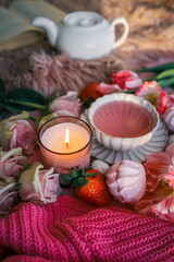 Flat lay with a pink tea cup, candle, tulips, roses, meringues, and strawberries on a soft pink background. Cozy, romantic setting with a warm, feminine aesthetic and open book