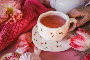 Cozy flat lay with pink tea in a floral cup, tulips, candlelight, sweets, and soft textures on a pink background. Feminine Valentine's or romantic morning mood