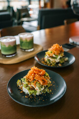 Close-up of a gourmet salad stack with creamy dressing and caramelized onions, served on a dark plate in a cozy modern café setting with blurred background