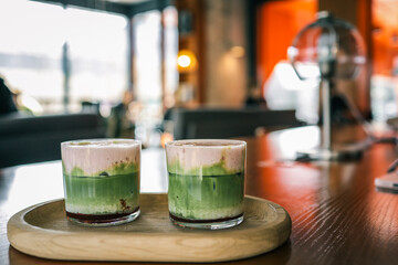 Two glasses of layered matcha and berry lattes served on a wooden tray in a cozy modern café with a blurred background of people and interior