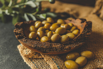 Rustic wooden bowl filled with green olives on burlap fabric with olive branch in background. Natural warm lighting and dark wood backdrop add to the organic Mediterranean atmosphere