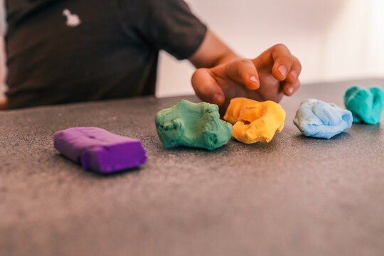 Child's hand playing with colorful modeling clay pieces on a gray table. Soft playdough in various colors including purple, green, yellow, and blue