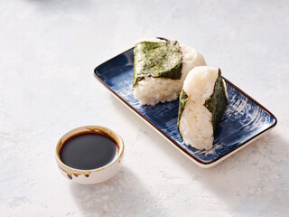 Two delicate onigiri rice balls wrapped in seaweed rest on a blue platter, accompanied by a small bowl of rich soy sauce, evoking a sense of culinary heritage in a peaceful ambiance