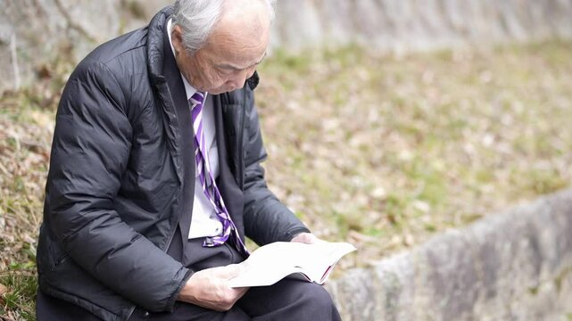 A winter park in Japan. A 78-year-old Japanese man in a suit and a down jacket sits on concrete. He reads white books.