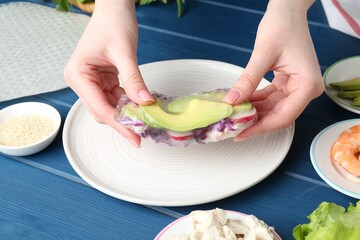 Woman making tasty spring roll at blue wooden table, closeup