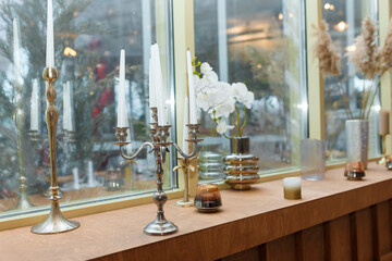Long white candles in candlesticks and vases with dry plants, artificial flowers on windowsill. Decorative elements in interior