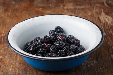 ripe fresh blackberries in white metal bowl on brown old rustic wooden table. Closeup shot