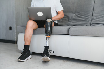 Full length portrait of adult man with prosthetic leg using laptop while sitting on sofa at home