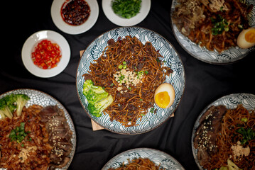 Flatlay angle fried ramen with chili oil served with beef beacon and broccoli also with half boiled egg on top. Served on a white and black pattern plate