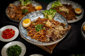 Landscape angle fried udon with chili oil served with sliced beef teriyaki and broccoli also with half boiled egg on top. Served on a white and black pattern plate