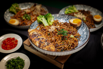 Landscape angle Fried ramen with chili oil served with sliced chicken charsiu and broccoli also with half boiled egg on top. Served on a white and black pattern plate