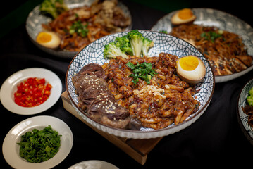 Landscape angle fried udon with chili oil served with beef barbeque and broccoli also with half boiled egg on top. Served on a white and black pattern plate.