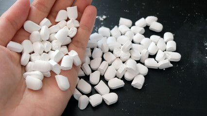 Hand with handful of white supplement tablets, crushed into half. With a big pile of the same tablets on the table, in the background.