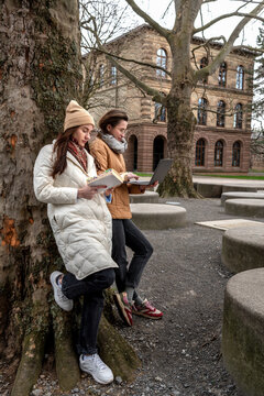 Two female students study outdoors, balancing laptops and books in the cool breeze