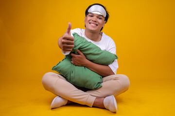 Asian Young Man in Pajamas Sitting While Holding Pillow and Showing Thumb Up