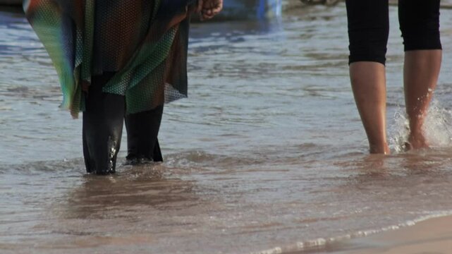 Woman wearing burkini walks barefoot in seawater along sandy beach. Swimsuit for Muslim girls who practice Islam. Close up shot of female legs. Two girlfriends walking on shore. Rear back view
