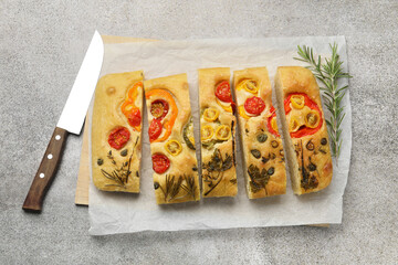 Cut delicious focaccia with flowers made of vegetables, knife and rosemary on grey table, top view