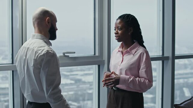 Medium shot of bicultural business partners in formalwear shaking hands standing by panoramic windows at office space