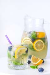 Chilled citrus drink with lemon slices, blueberries, mint, and ice in a glass and jug, on a white background.