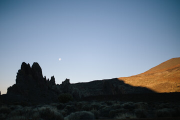 A dramatic landscape on the way to a volcano, featuring rocky terrain under a clear blue sky. The moon is visible in the daytime sky, and the scene is divided between shadowed and sunlit sides.