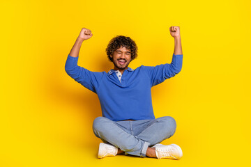 Young stylish man in casual outfit sitting on the yellow background, raising hands in excitement,...
