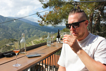young man in sunglasses drinking and tasting wine, brandy and water in the garden with an overview on vineyards in Tsiakkas Winery in Cyprus on a sunny day